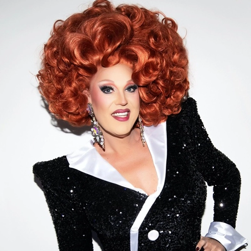 A person with a large, curly, bright red wig smiles at the camera. Channeling Dixie Krystals, they wear dramatic makeup, jeweled earrings, and a sparkly black sequined blazer with bold white lapels and a large button, standing against a plain background.