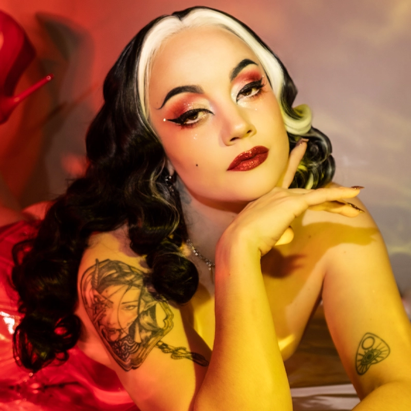 A person with long, wavy black hair featuring a dramatic white streak strikes a pose reminiscent of Logan L'Enfer, hand under chin. Bold red lipstick, winged eyeliner, shimmery eyeshadow, and arm tattoos shine in the warm red and yellow lighting.