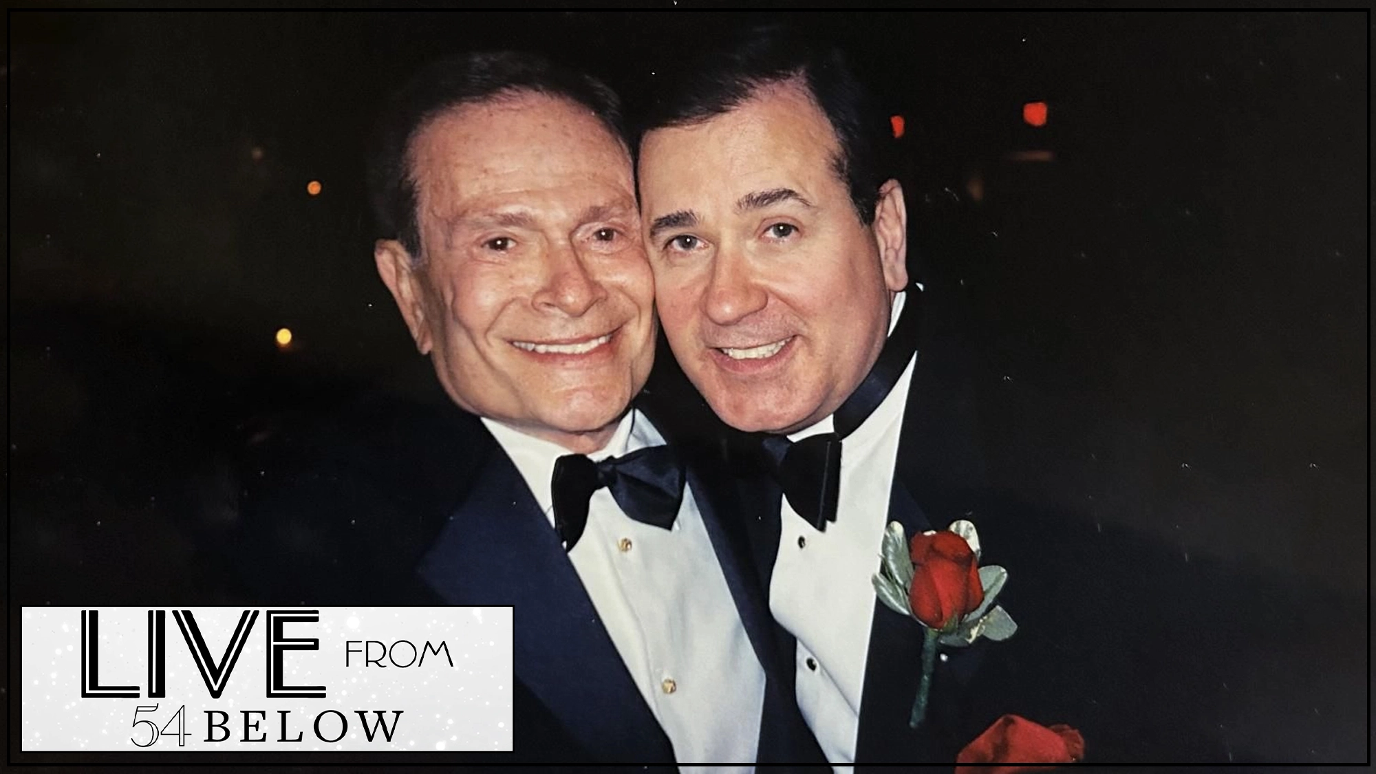 Two smiling men in tuxedos, including Lee Roy Reams, pose together at an event. One wears a red rose boutonnière. The dimly lit background glows softly, and a 