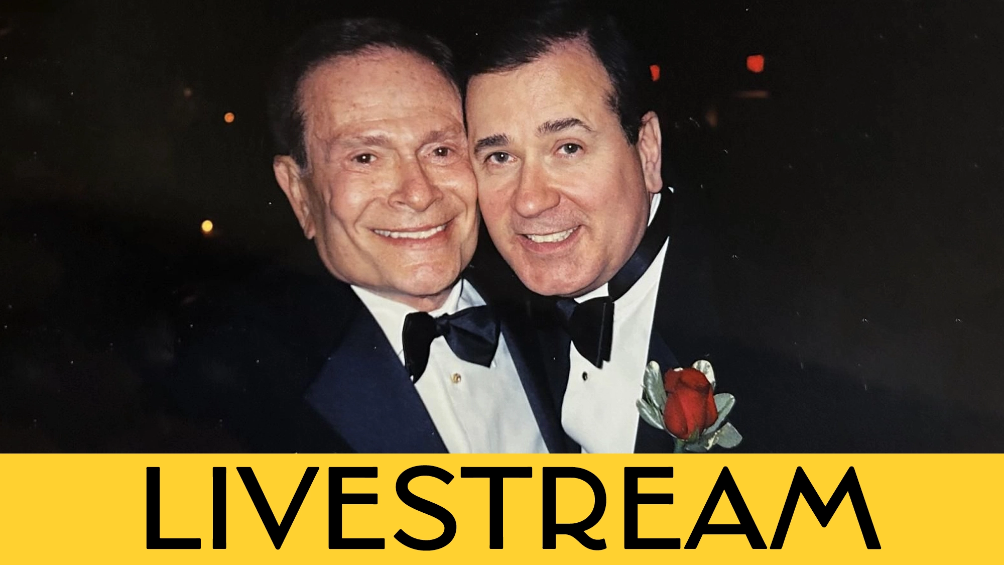 Two older men, Jerry Herman and Lee Roy Reams, smile and embrace in tuxedos. One wears a red rose boutonnière. The background is dark and blurry. A yellow banner at the bottom reads 