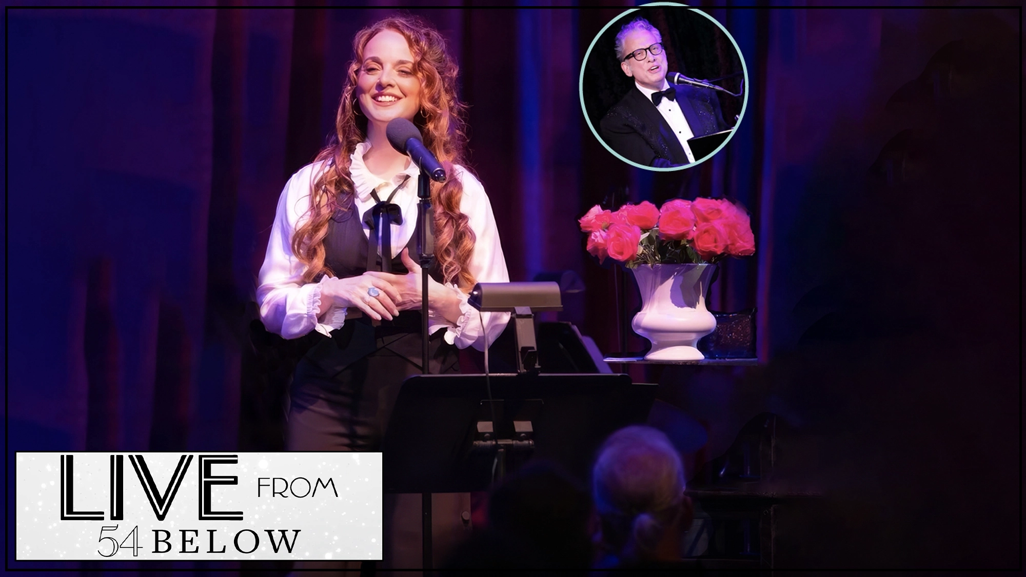 A woman with long, wavy red hair stands at a microphone on stage, smiling, wearing a black vest over a white shirt. 