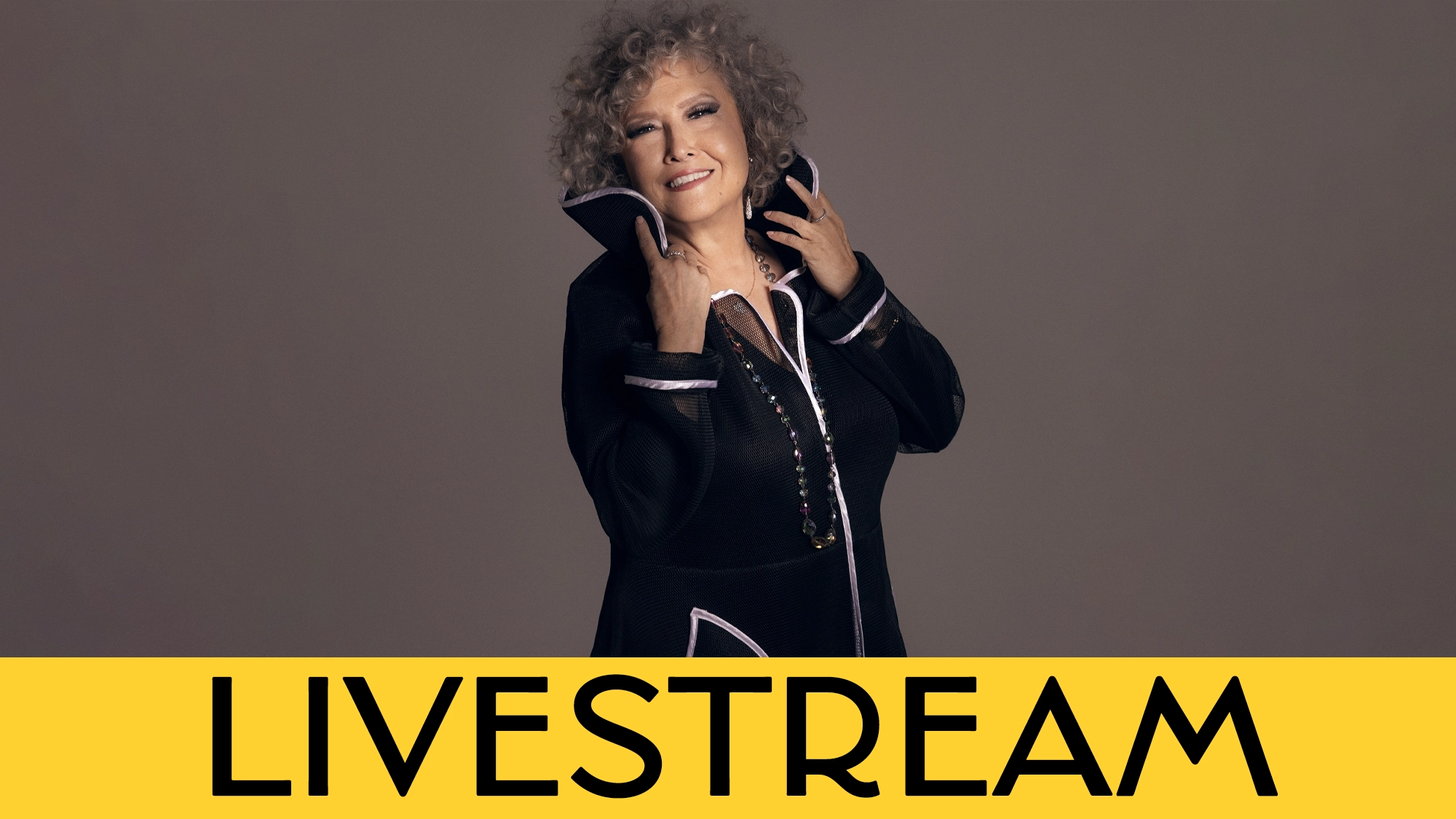 A smiling woman with curly gray hair stands against a plain background, wearing a black outfit with a high collar and necklace. She touches her collar with both hands. A yellow banner at the bottom reads 