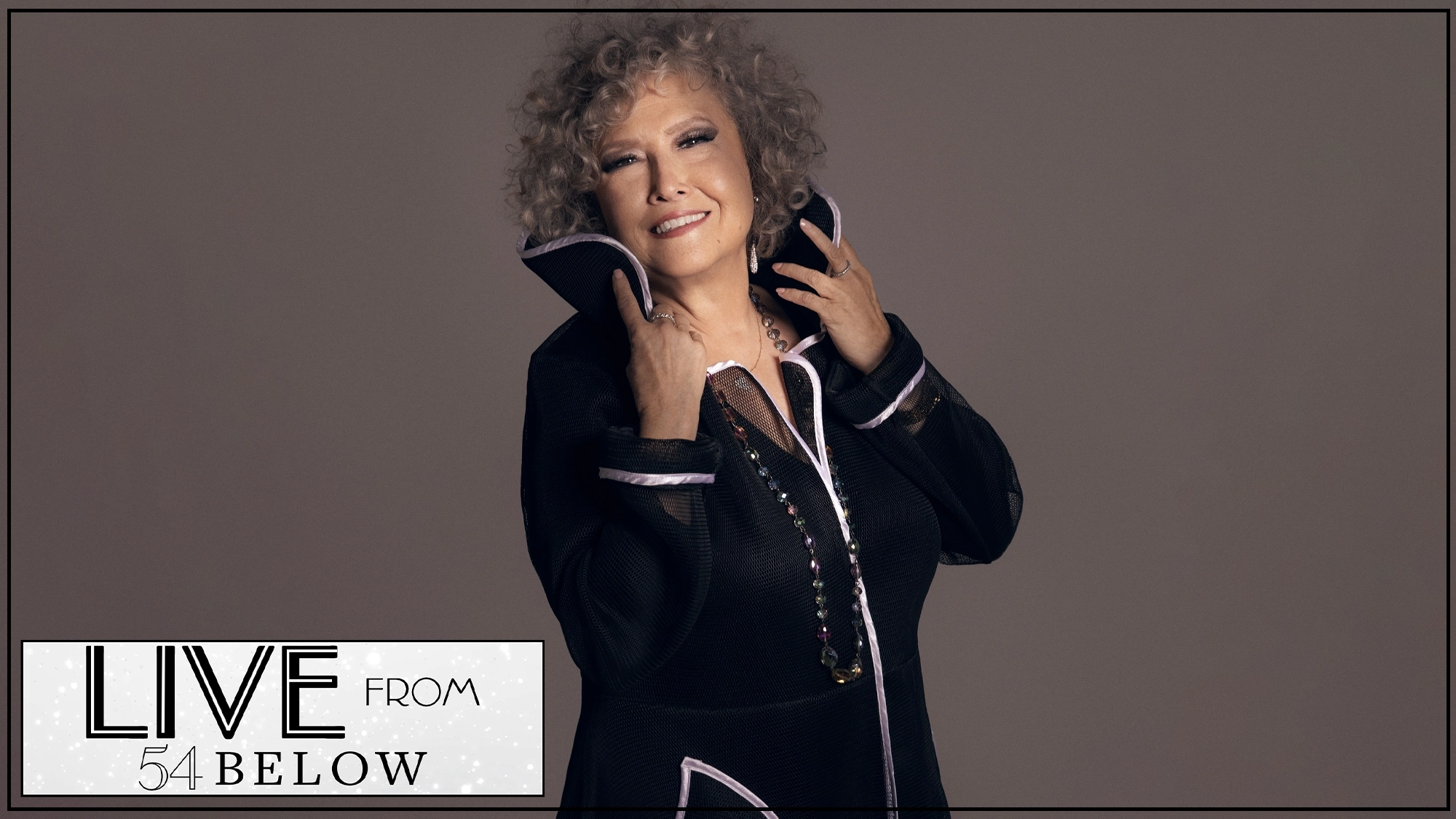 A smiling older woman with curly gray hair poses in a stylish black outfit with a high collar. She holds the collar with both hands. The background is neutral. “LIVE FROM 54 BELOW” and 