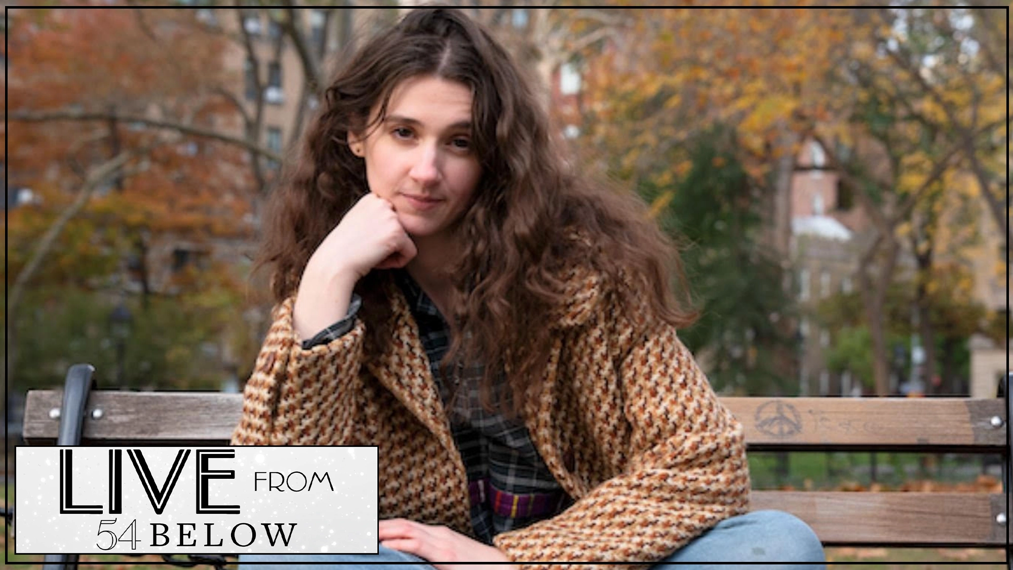 A person with long wavy brown hair sits on a park bench, resting their chin on their hand and smiling slightly. They wear a brown checkered coat and blue jeans. Autumn trees fill the background. The LIVESTREAM “Live from 54 Below” logo appears in the corner.
