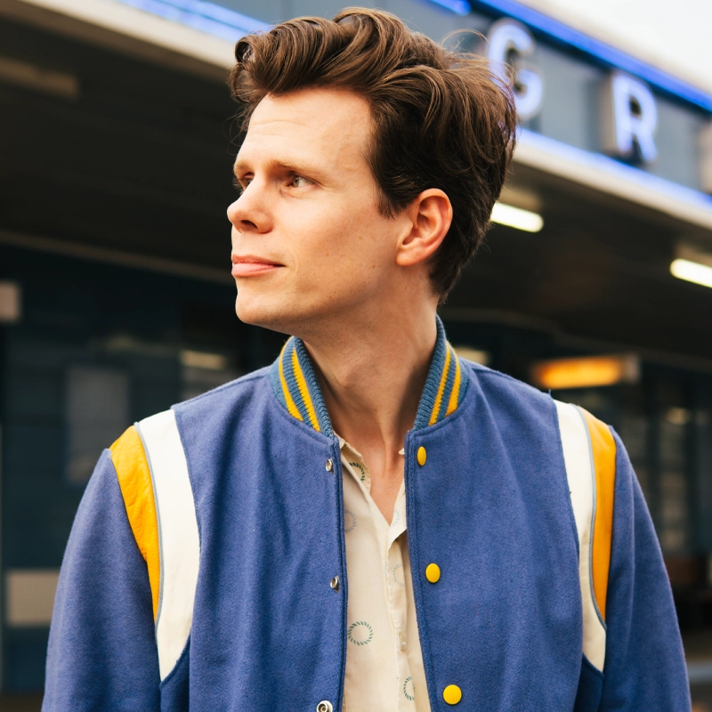 A young man with styled brown hair wears a blue varsity jacket with yellow and white stripes. He looks to his left with a slight smile, standing outside in front of a building with blue walls and large white letters partially visible above him.