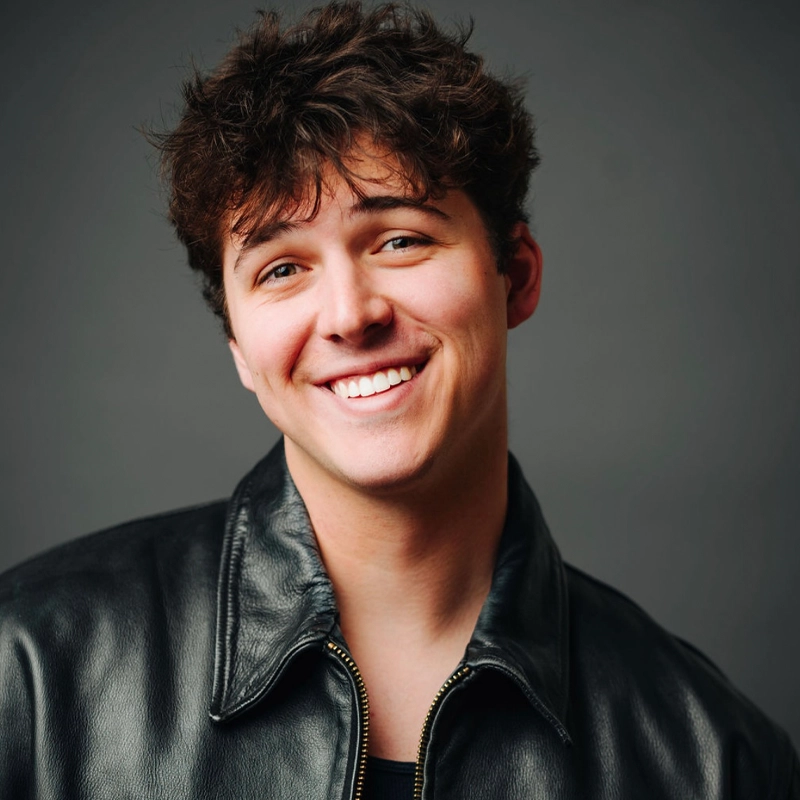 A young man with wavy brown hair smiles warmly at the camera. He wears a black leather jacket over a dark shirt. The background is plain and gray, keeping the focus on his friendly expression and casual appearance.