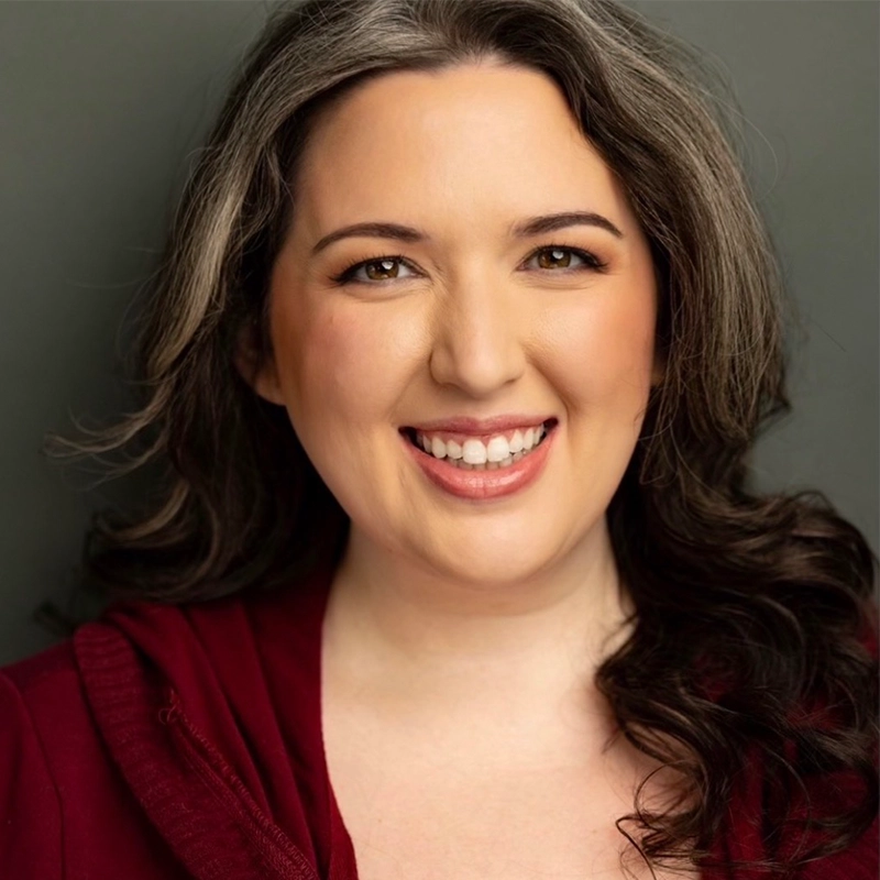 Jett Roberts, a woman with long wavy dark hair and gray highlights, smiles warmly at the camera. She is wearing a burgundy top and natural makeup, with a neutral background behind her.