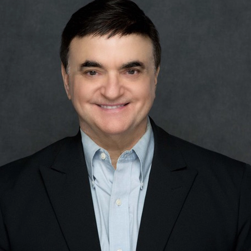 A middle-aged man with short dark hair, identified as Lawrence Leritz, is smiling at the camera. He is wearing a dark suit jacket over a light blue collared shirt, set against a plain, dark gray background for a professional portrait feel.