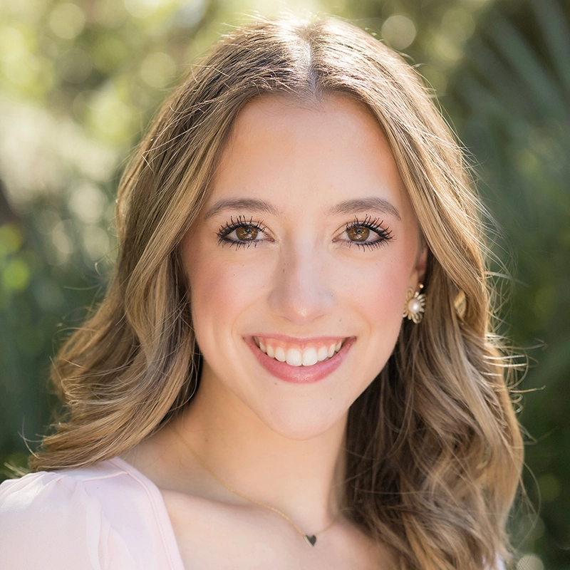 A young woman with wavy blonde hair smiles at the camera. She wears a light pink top, a delicate gold necklace, and floral stud earrings from Danté X Johnson. The background is blurred greenery, giving a soft, natural outdoor feel.