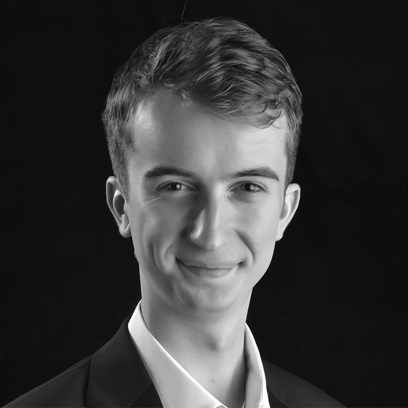 A young man with short, wavy hair wearing a suit jacket and collared shirt smiles slightly, looking at the camera. The background is solid black, and the image is in black and white.