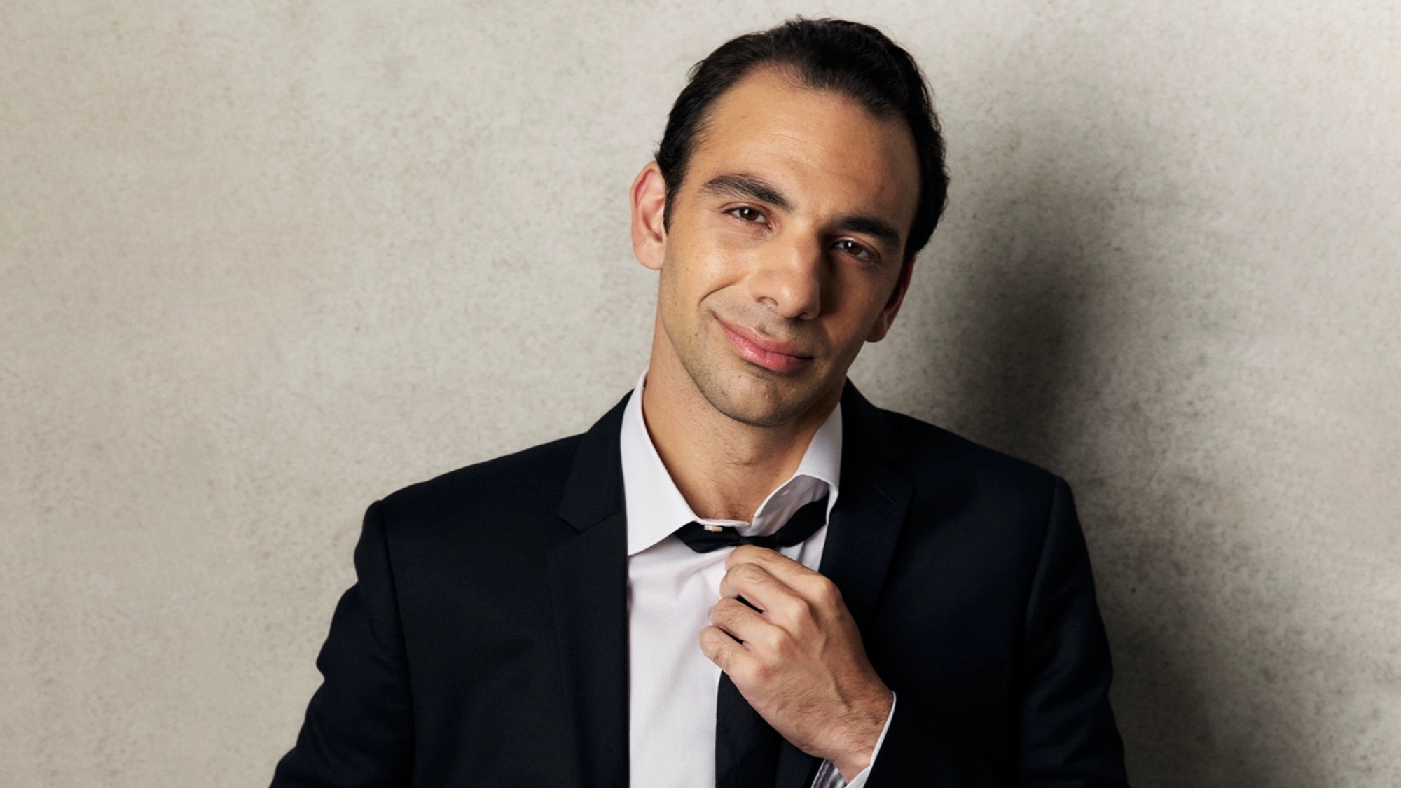 A man in a dark suit and white shirt stands against a light, textured wall. Channeling Martin Bonventre’s Swingin’ in a New Dimension, he looks at the camera with a slight smile and adjusts his loosened black tie, appearing relaxed and confident.