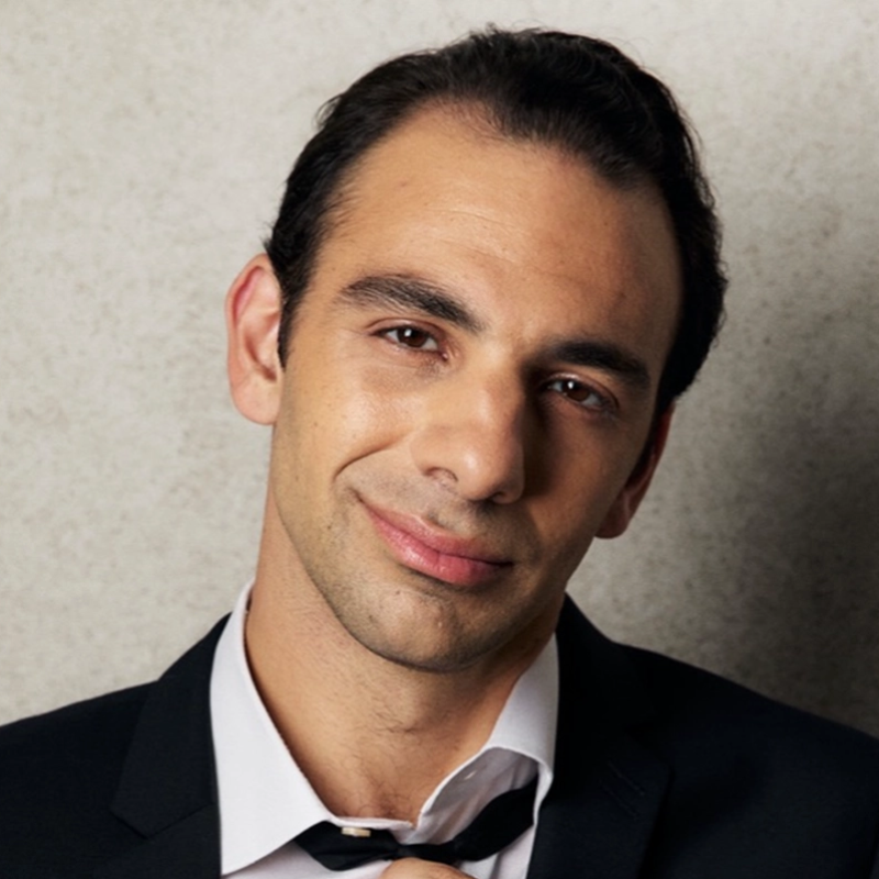A man with short dark hair and brown eyes wears a black suit jacket and white shirt with the collar slightly open. He smiles gently, tilting his head slightly, with a loosely knotted black tie. The background is a light, textured surface.