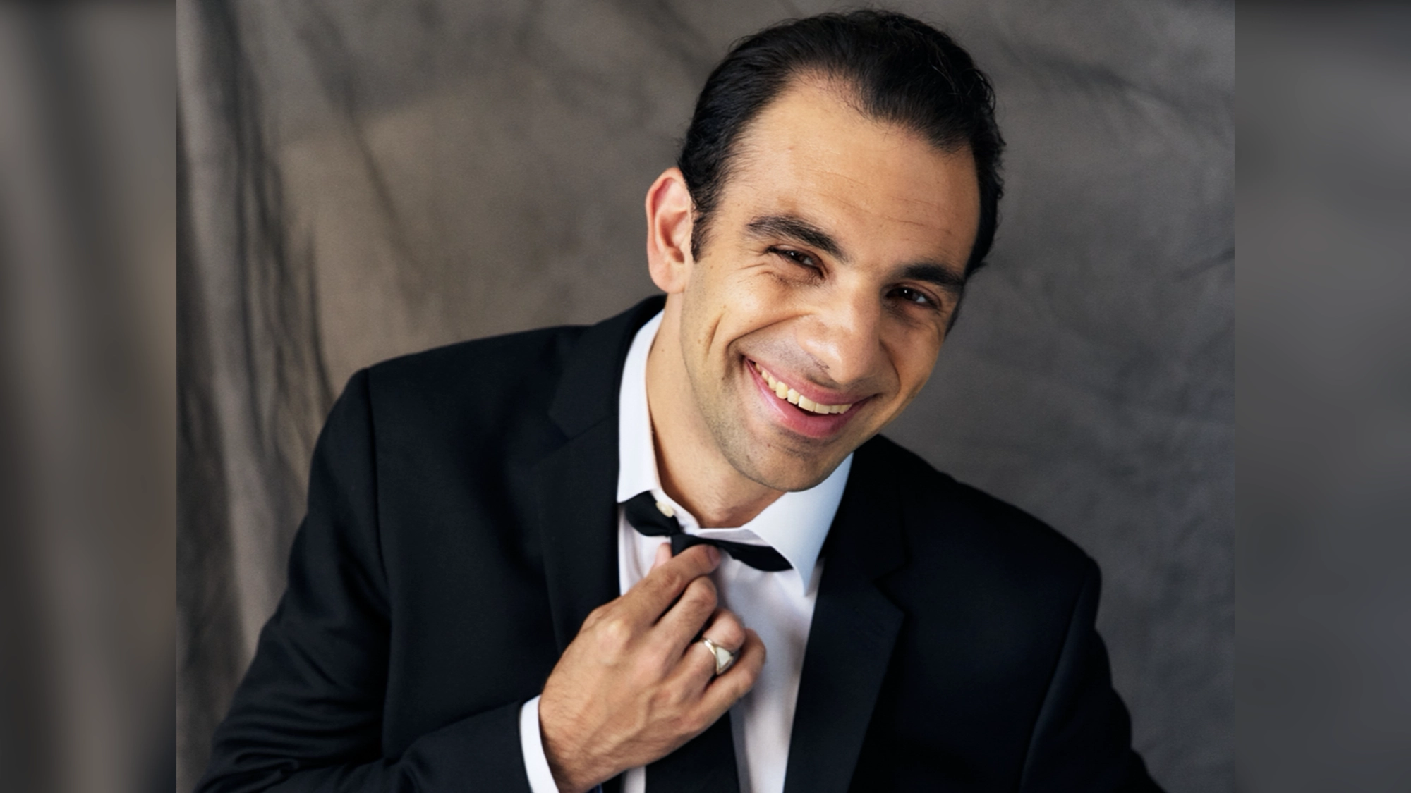 Martin Bonventre smiles warmly at the camera, adjusting his black tie with his right hand. Dressed in a sharp suit, he stands against a soft gray background—capturing the charm of Swingin’ in a New Dimension.