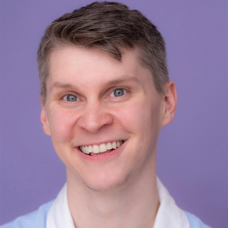 A young adult with short, light brown hair and blue eyes smiles widely at the camera. They are wearing a light blue collared shirt over a white top. The background is solid light purple.