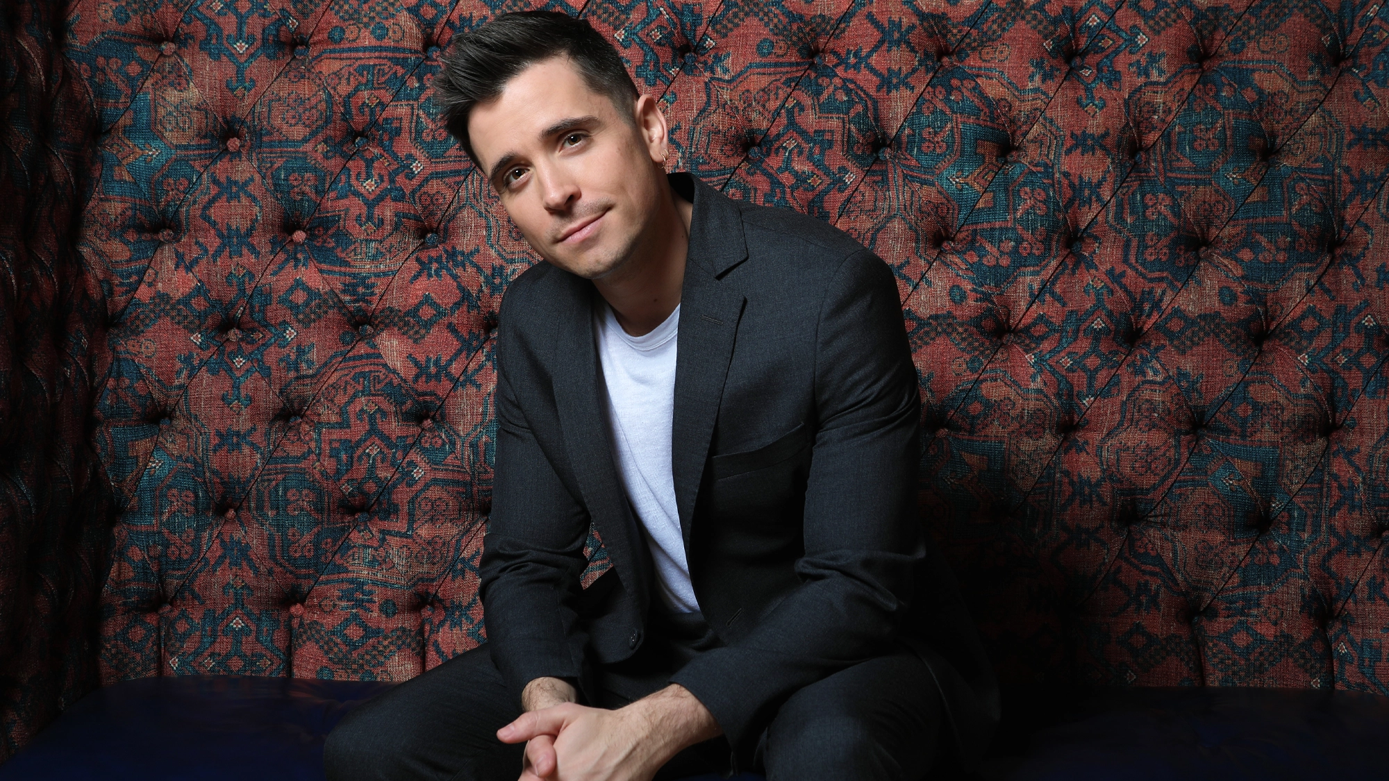 Matt Doyle - Site Image Matt Doyle, with short dark hair, wearing a dark gray suit and white t-shirt, sits leaning forward with hands clasped. He smiles slightly and poses against a tufted, ornate red and blue patterned background to Make The Season Bright.