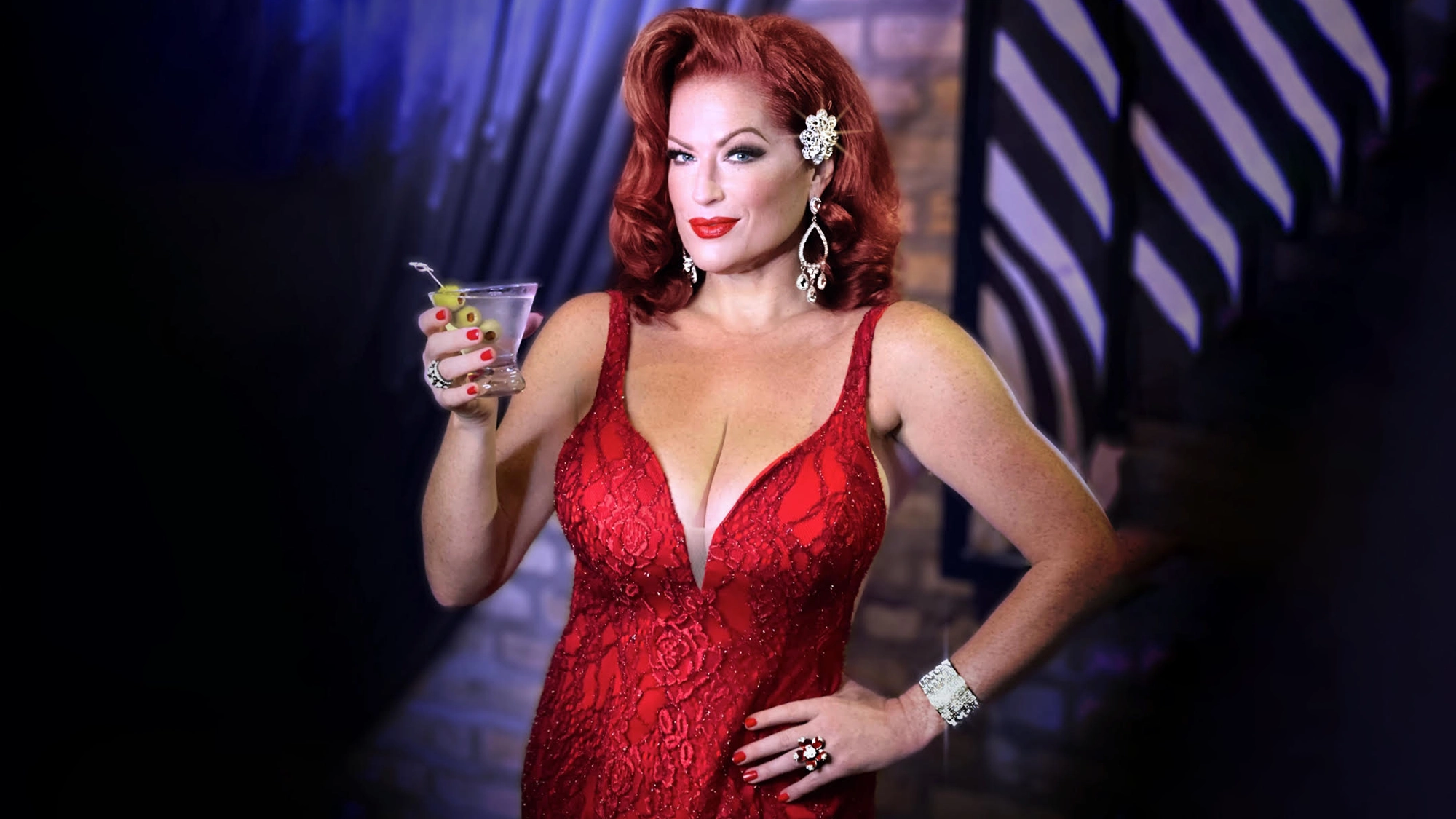 A glamorous person with wavy red hair in a red, low-cut lace dress holds a cocktail and poses confidently at GAYCON. They wear bold red lipstick, large earrings, a jeweled hairpiece, bangles, and rings against a dark background of blurred geometric patterns.