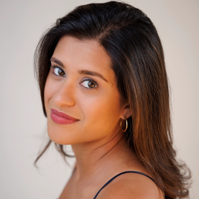 Francesca Mehrotra, with medium-length dark brown hair and light brown skin, looks over her shoulder, smiling softly. She has brown eyes, gold hoop earrings, and a thin-strap top. The softly lit background highlights her face.