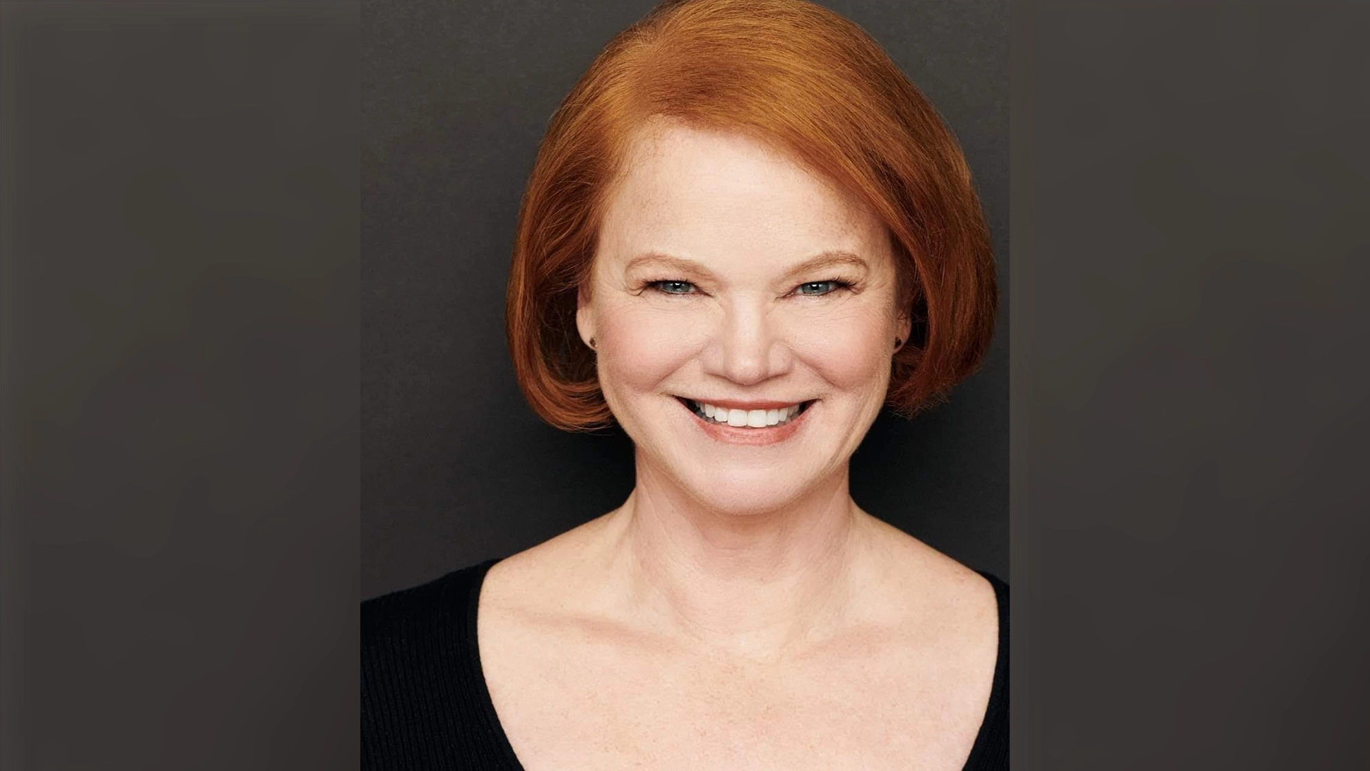 Melodie Wolford, with short red hair and fair skin, smiles at the camera in a black scoop-neck top. The bright lighting highlights her features against a solid dark grey background, as if she’s Waiting In The Wings for her next moment.