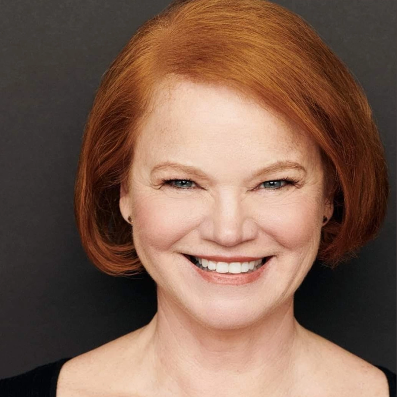 A woman with fair skin, straight shoulder-length red hair, and blue eyes is smiling warmly. She is wearing a black top and is posed against a plain dark background. Her teeth are visible, and she has light makeup and small stud earrings.