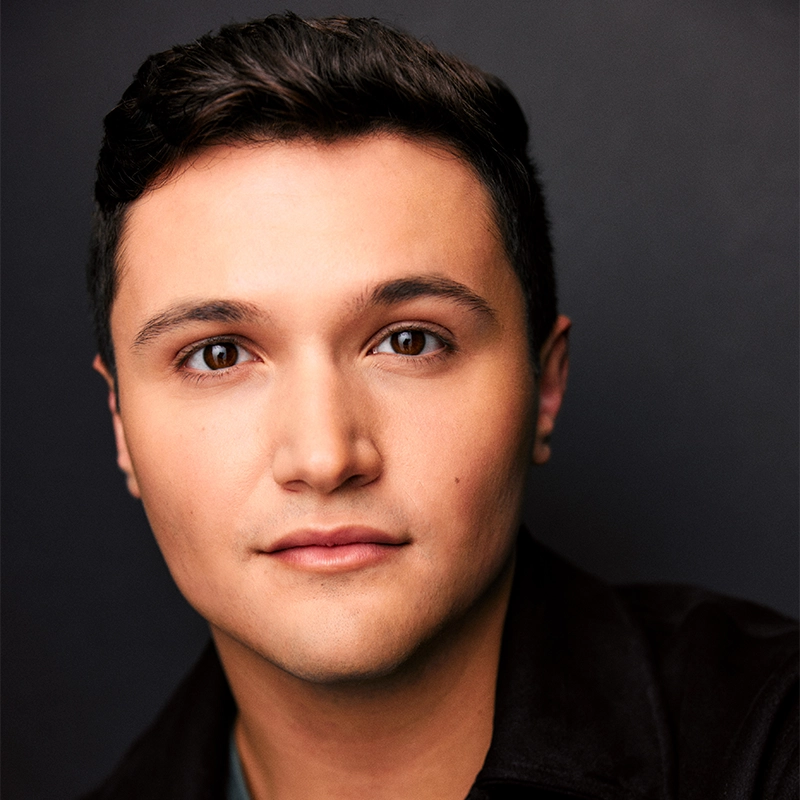 A young man with short, dark hair and brown eyes looks directly at the camera with a neutral expression. He has clear skin and smooth features, wearing a dark jacket over a green shirt against a plain dark background.