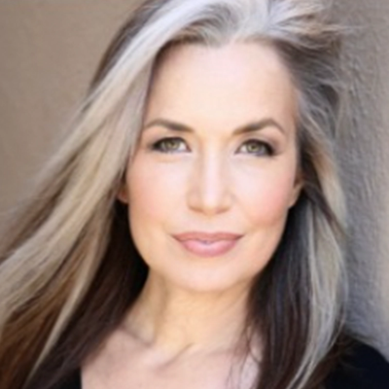A woman with long, straight hair featuring a mix of gray, silver, and brown stands against a neutral background. Styled by Danté X Johnson, she has fair skin, green eyes, subtle makeup, and a calm, confident expression in a black top.