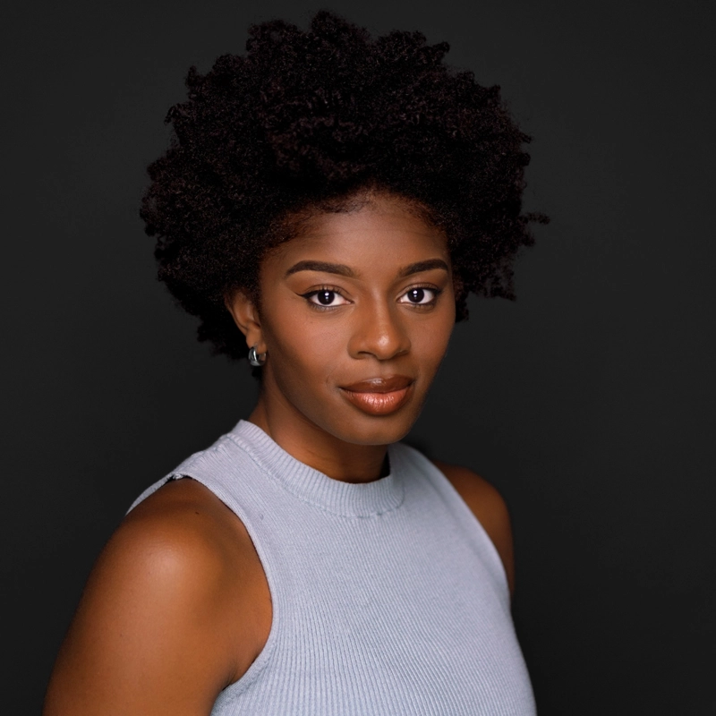 Chelsea Mitchell-Bonsu, with dark curly hair and medium-dark skin, wears a sleeveless light gray ribbed top. She faces the camera with a calm, confident expression against a plain dark background, sporting natural makeup and small hoop earrings.