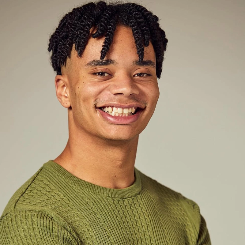 Alejandro MullerDahlberg, a young man with short, twisted black hair, smiles at the camera. He wears a light olive green textured sweater and stands against a softly-lit beige background, arms loosely crossed and body slightly angled.