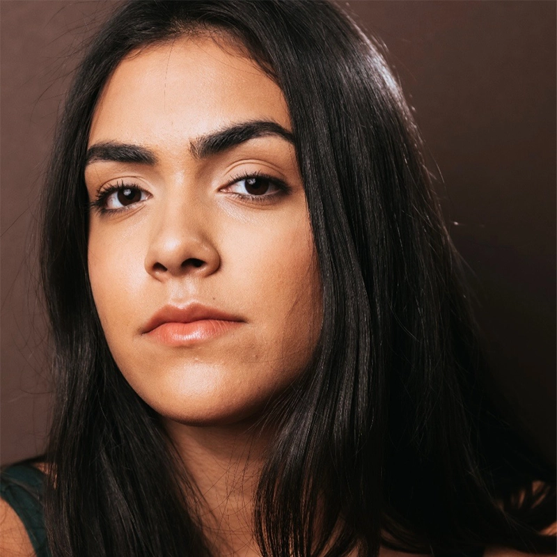 Annie Thomas, a young woman with long dark hair and thick eyebrows, gazes confidently at the camera. Her smooth skin and natural makeup stand out against the plain, dark background, drawing focus to her striking features.