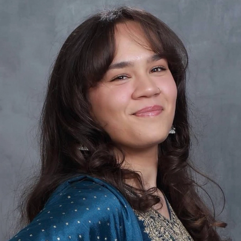 Jett Roberts, with long, wavy brown hair, smiles softly while posing against a gray background. Wearing a blue and gold traditional outfit with intricate detailing and small, dangling earrings, the atmosphere is calm and confident.