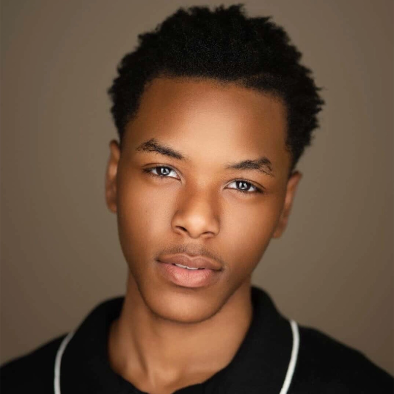 Samuel Nelson III, a young man with short, textured black hair and smooth skin, looks directly at the camera. He wears a black collared shirt with white trim, set against a soft, warm brown background with lighting that highlights his features.