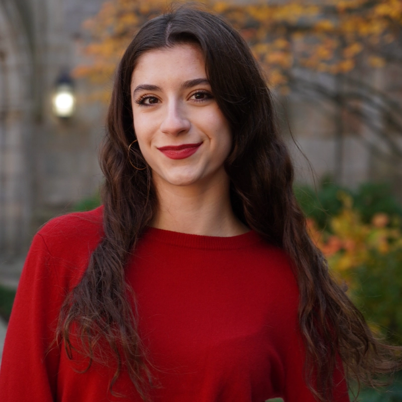 A young woman with long wavy brown hair and red lipstick smiles at the camera. Iara Nemirovsky is wearing a red sweater and standing outdoors, with blurred autumn foliage and stone architecture in the background.