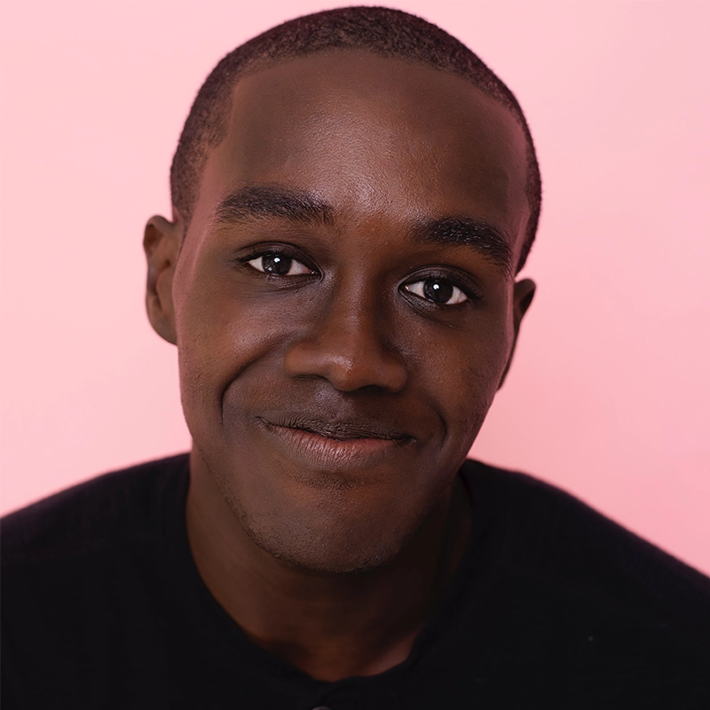 A young man with dark skin and short hair smiles gently. He is wearing a black shirt and is posed against a soft pink background, looking directly at the camera in this close-up portrait by Audrey Graves.