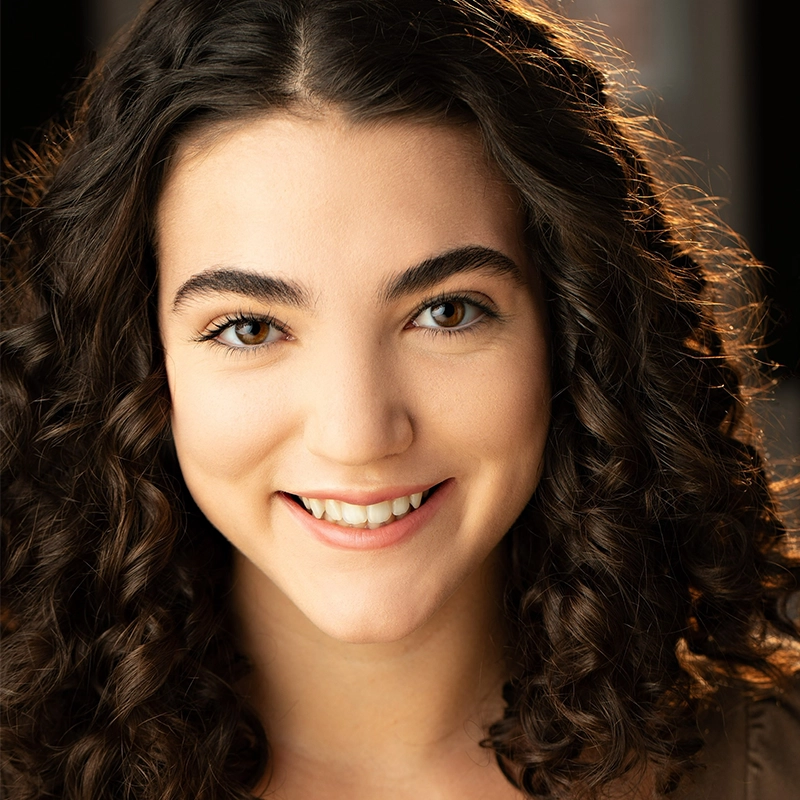 A young woman with medium skin, dark curly hair, and thick eyebrows smiles warmly at the camera. Lit by natural sunlight, she exudes a soft, inviting atmosphere. The softly blurred background adds depth to this Danté X Johnson-inspired portrait.