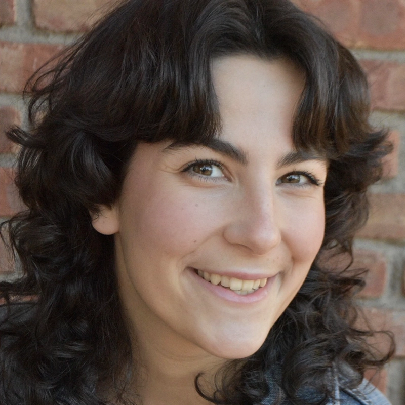 Jett Roberts, a young person with wavy dark hair and fair skin, smiles at the camera in front of a brick wall. They have brown eyes, visible eyelashes, and wear a denim jacket under soft, natural lighting.