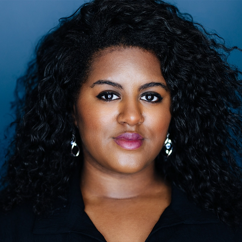 Praise Oranika, a woman with medium-dark skin and long, curly black hair, poses confidently in front of a blue background. She wears drop earrings, a black top, and natural makeup with subtle eyeliner and pink lipstick.