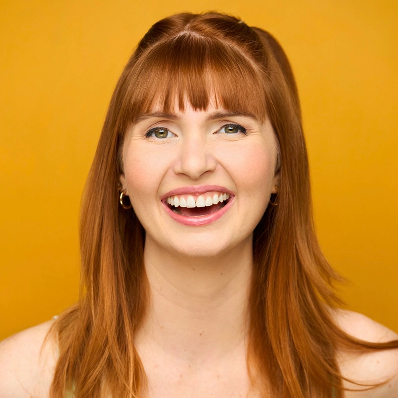 A young woman with straight, long red hair and bangs smiles brightly, showing her teeth. Jett Roberts wears gold hoop earrings and a light top. The vibrant yellow background creates a cheerful and warm atmosphere.