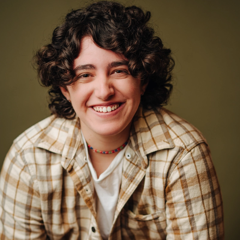 Cameron Parker, with curly dark hair, smiles warmly at the camera. They wear a beige and brown plaid shirt over a white T-shirt and a colorful beaded necklace, set against a plain olive green background that creates a soft, inviting atmosphere.