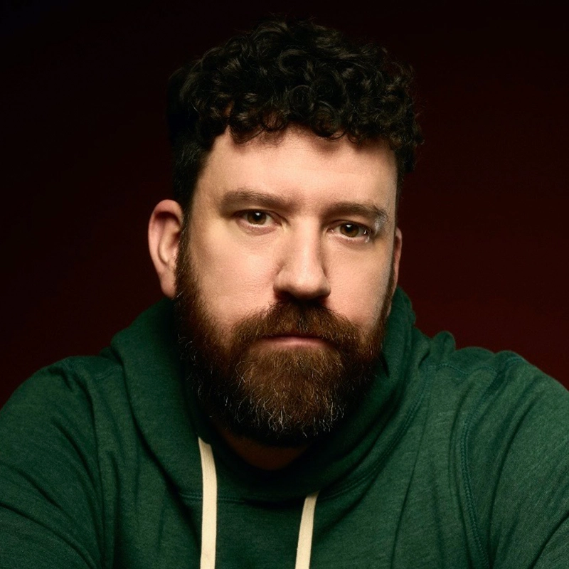 A man with curly dark hair and a full beard, identified as Justin Fischer, wears a green hoodie with white drawstrings. He sits against a dark red background, looking directly at the camera with a neutral expression as light highlights his face.