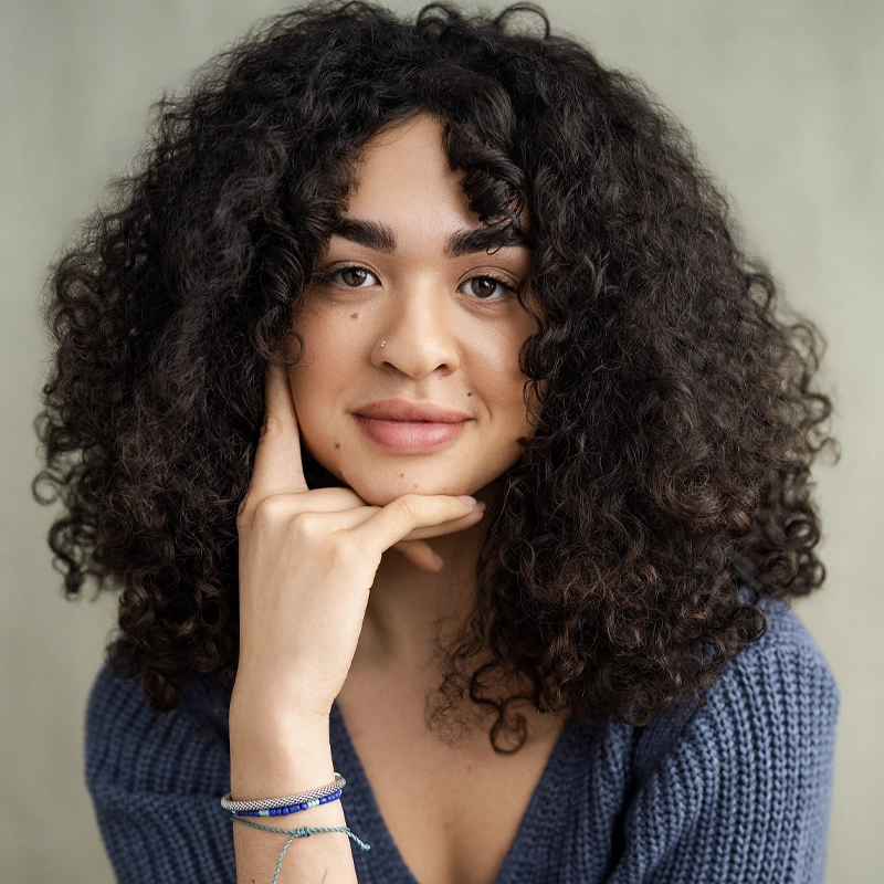 A young person with medium skin, curly dark hair, and a nose piercing smiles gently—a look reminiscent of Ilia Isorelýs Paulino. They wear a textured blue sweater, resting their chin on their hand against a softly blurred neutral backdrop.