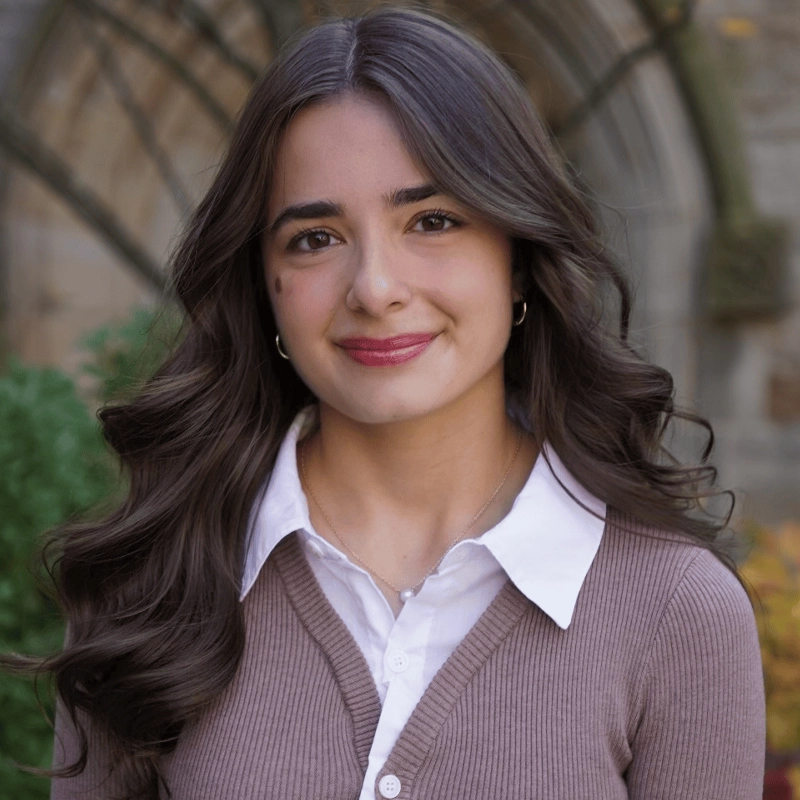 Gabby Pavlov, a young woman with long, wavy brown hair and a prominent beauty mark on her left cheek, smiles softly. She wears a white collared shirt under a taupe sweater, standing before a blurred stone archway and lush greenery.