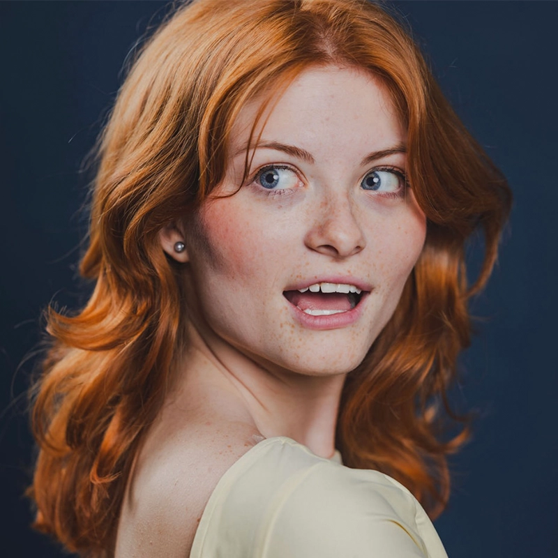 A young woman, Piper Redford, with wavy red hair and fair, freckled skin looks over her shoulder, mouth slightly open in surprise or delight. She wears a light-colored top and small earrings, set against a dark blue background.