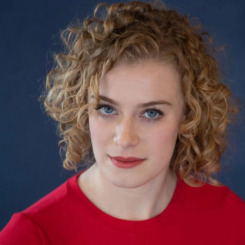 A woman with fair skin and curly blonde hair looks directly at the camera. She has blue eyes and wears a red top. The background is a solid dark blue, and the lighting highlights her facial features clearly.