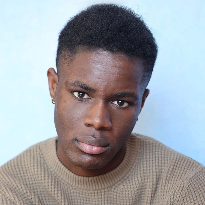 A young man with short, curly black hair and brown skin wears a beige knit sweater and small hoop earrings. Praise Oranika looks directly at the camera with a neutral expression, set against a softly blurred, light blue background.