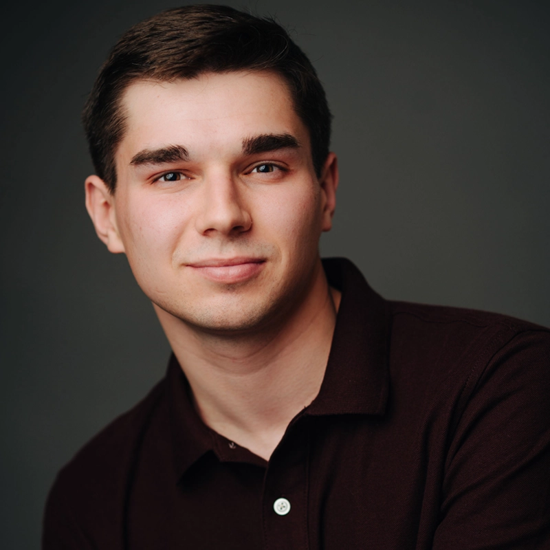 A young man with short brown hair and fair skin wears a dark maroon collared shirt. He smiles softly and looks directly at the camera. The background is plain and dark, giving the image a professional, studio-lit appearance.