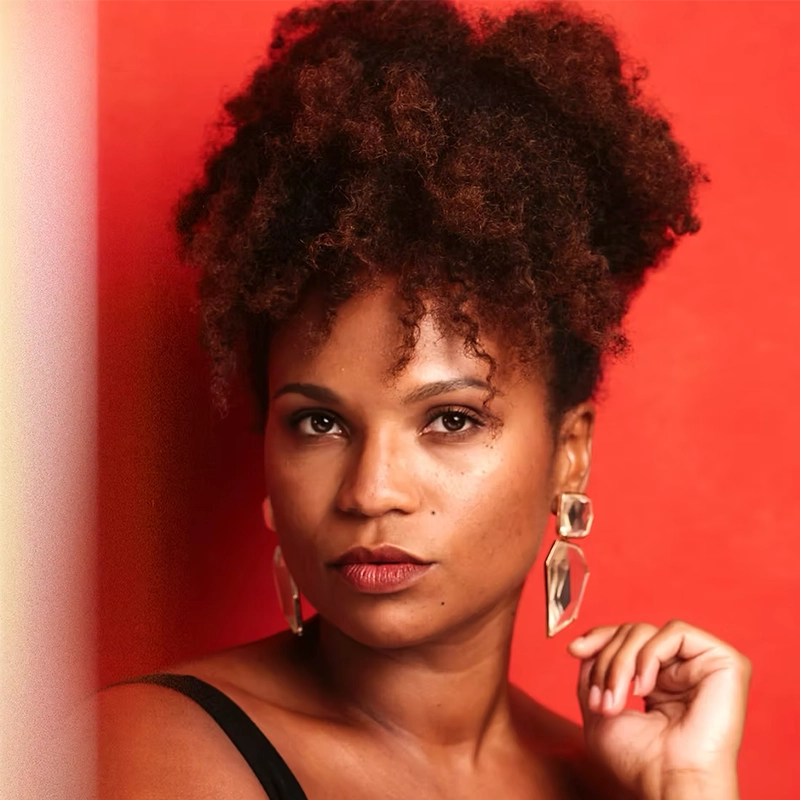 A woman with medium-brown skin and natural curly hair styled in a high puff poses against a vibrant red background. She wears St. Hubert-inspired geometric earrings shaped like clover, a black top, and bold lipstick. Her gaze is confident and direct.