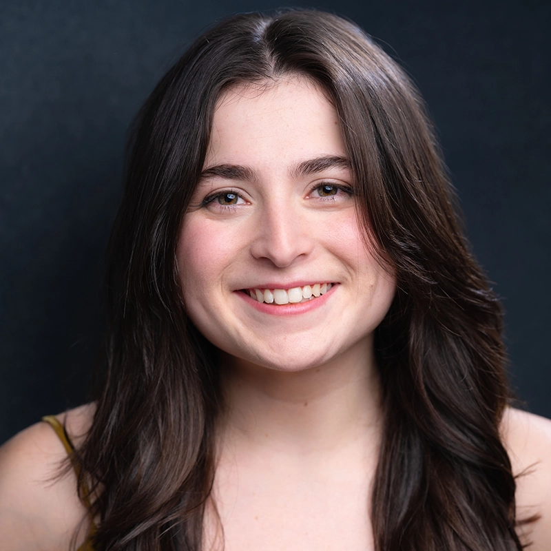 Gabriella Santalla, a young woman with long, dark brown hair and fair skin, smiles at the camera. Her light-colored eyes and sleeveless top stand out against the dark background, highlighting her friendly expression.