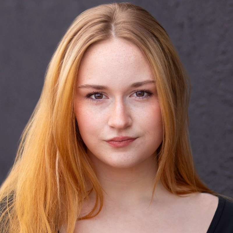 A young woman with long, straight, light brown hair and fair skin looks directly at the camera with a neutral expression. Jett Roberts is wearing a black top and stands in front of a dark, plain background.