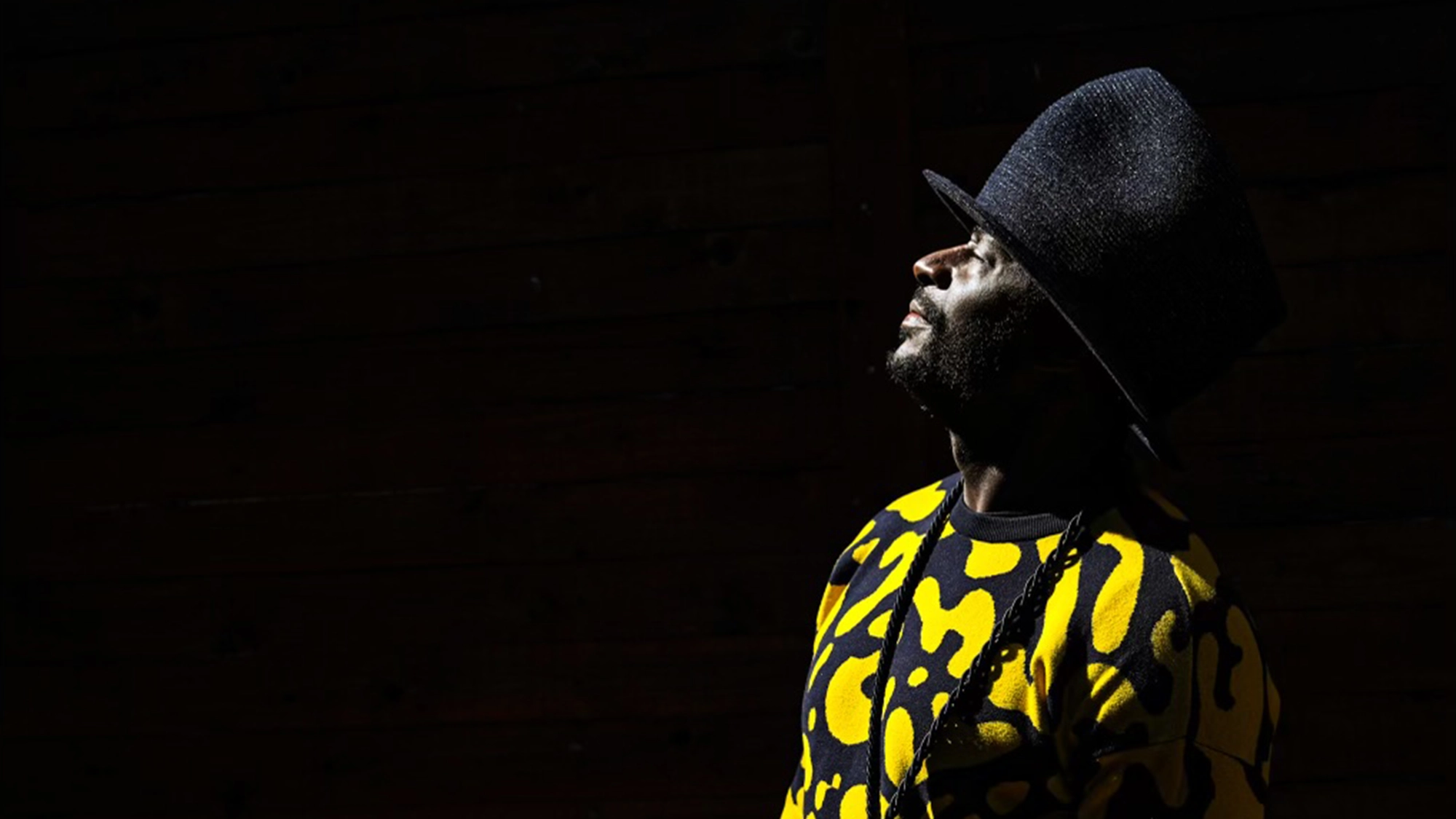 A man in a black wide-brimmed hat and a bright yellow and black patterned shirt stands in profile, looking upward into a beam of light. The background is dark, making the man’s face and clothing stand out vividly against the shadow.