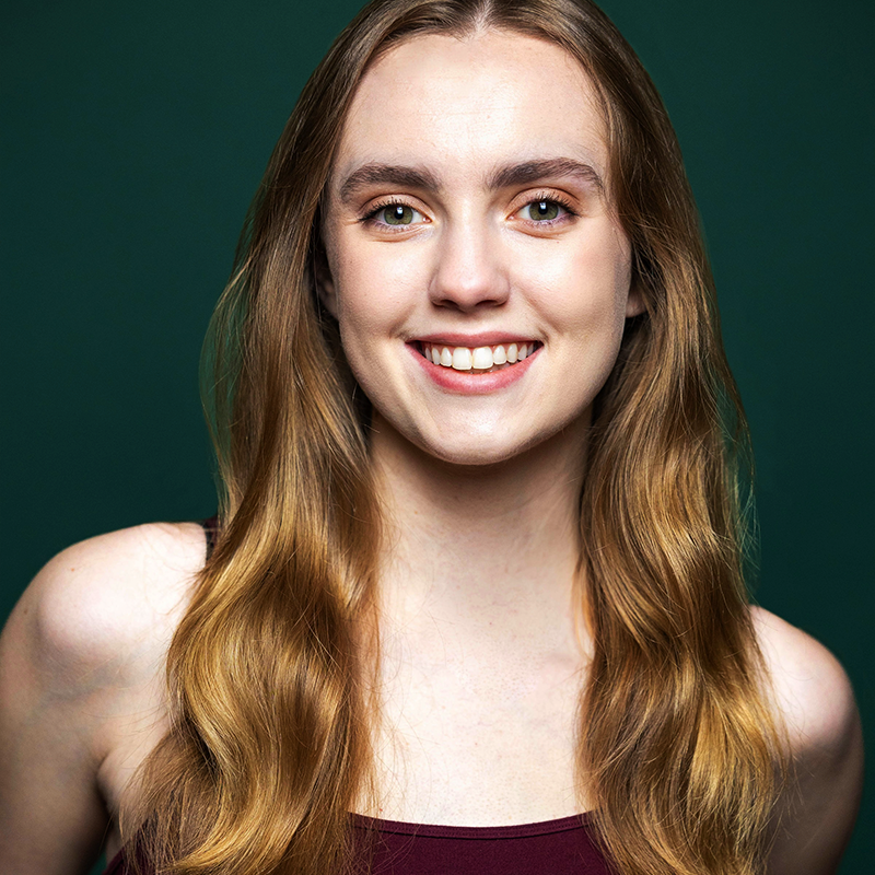 Stephanie Reed, a young woman with long, wavy, light brown hair, smiles warmly in a sleeveless burgundy top against a solid dark green background. Her expression is friendly and welcoming as she looks directly at the camera.