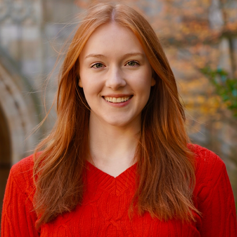 Lizzie Rowan, a young woman with long straight red hair and fair skin, smiles at the camera in a bright red sweater. The background is softly blurred, featuring stone architecture and autumn-colored foliage.